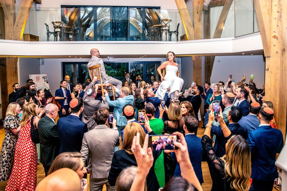 Bride and groom lifted by guests during lively wedding hora dance celebration in barn venue.