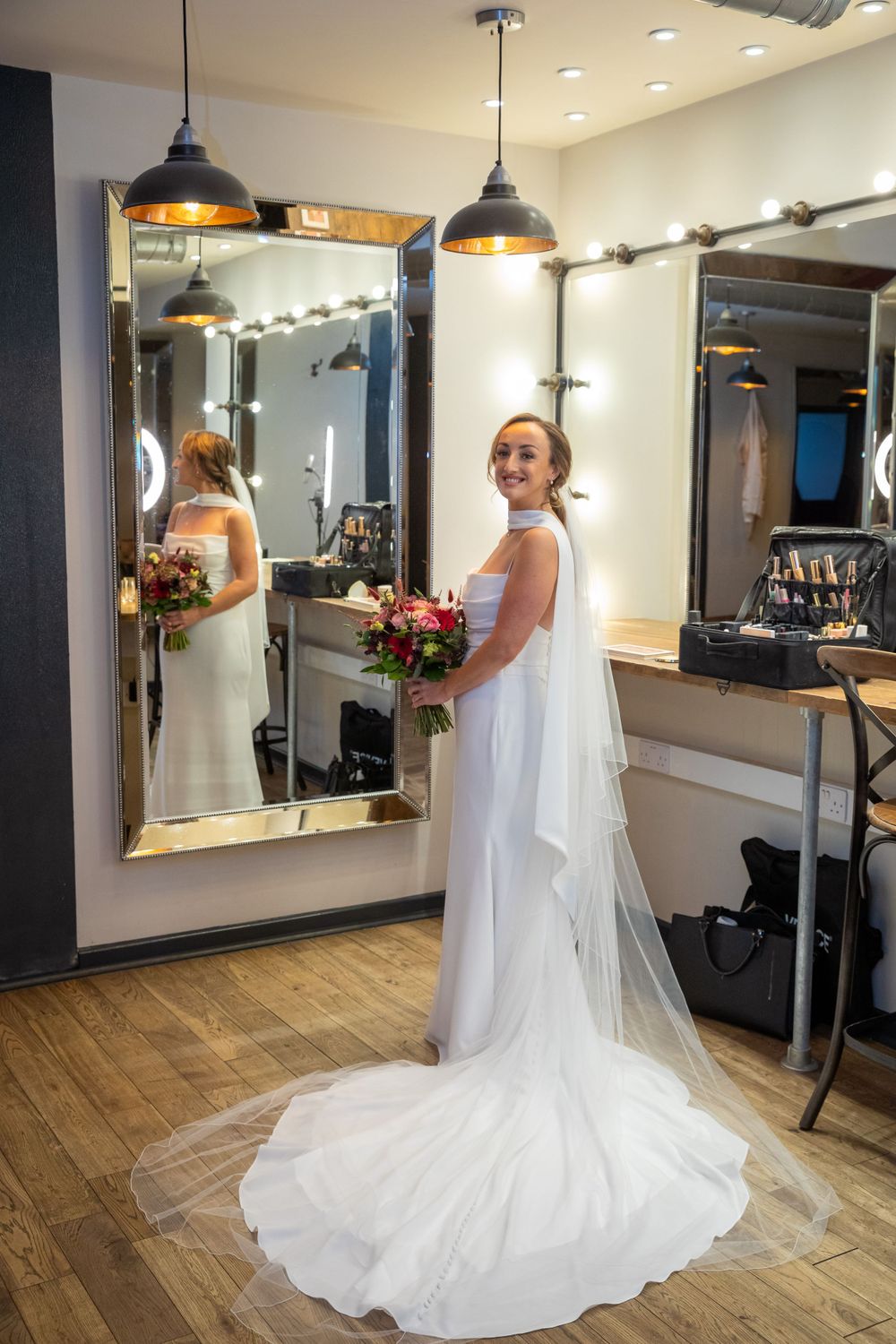 Bride in white gown holding bouquet stands before mirror in bridal suite on her wedding day.