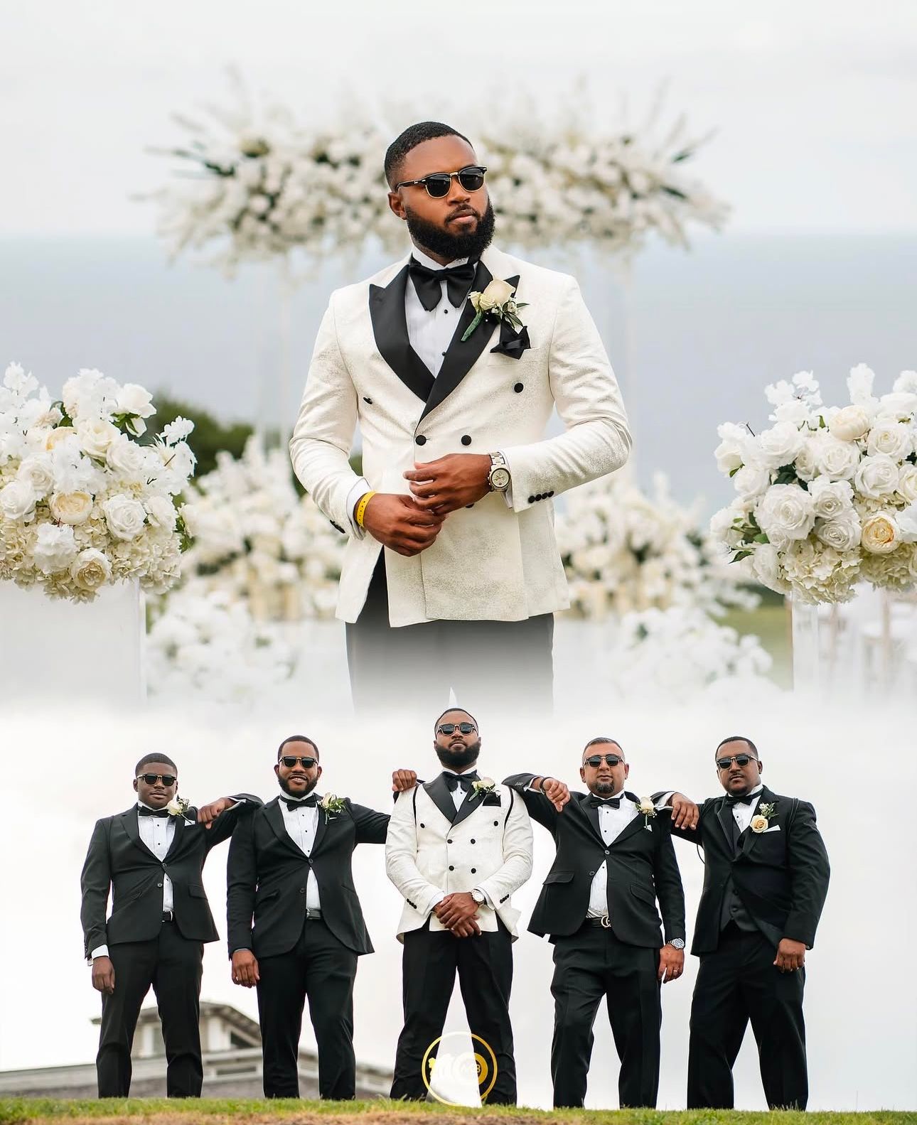 A wedding party in black tuxedos and one in white poses together outdoors with blooming white flowers in the background.