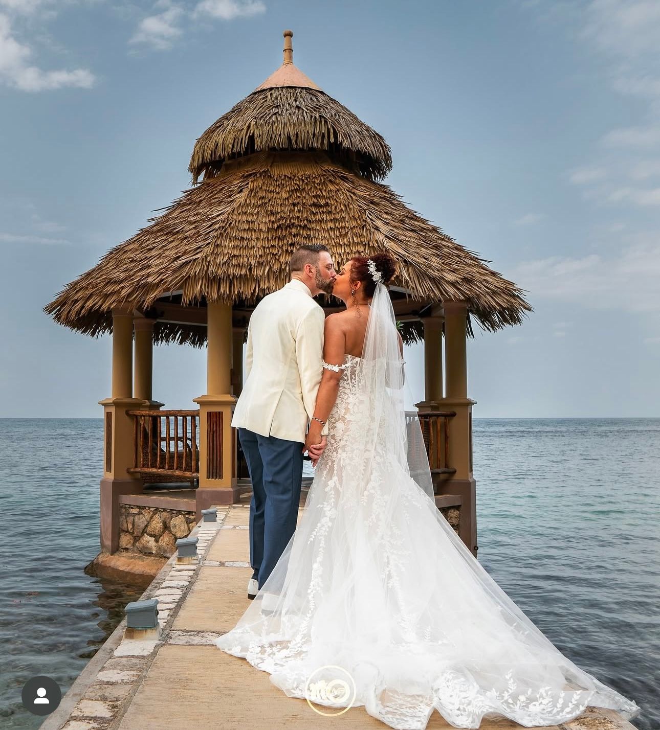 Wedding couple poses on wooden pier with thatched-roof gazebos overlooking ocean.