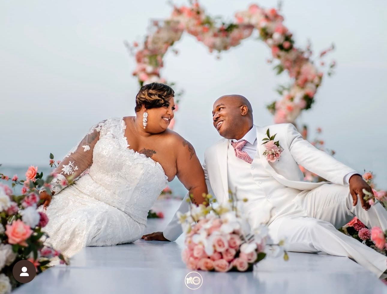 Wedding couple poses playfully on white backdrop with pink floral decorations.