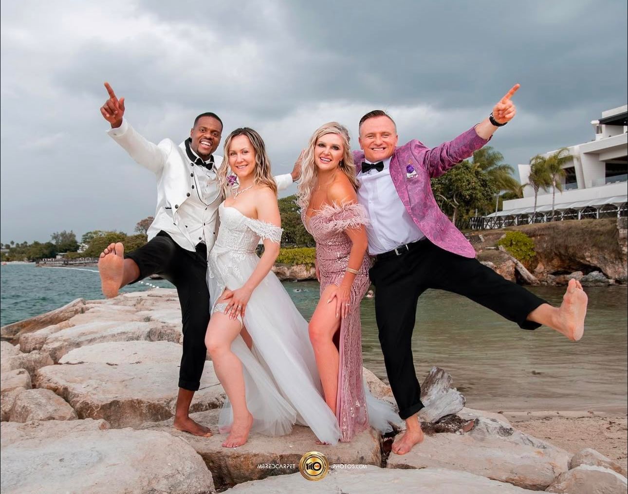 Wedding party poses playfully on rocky beach in formal attire.