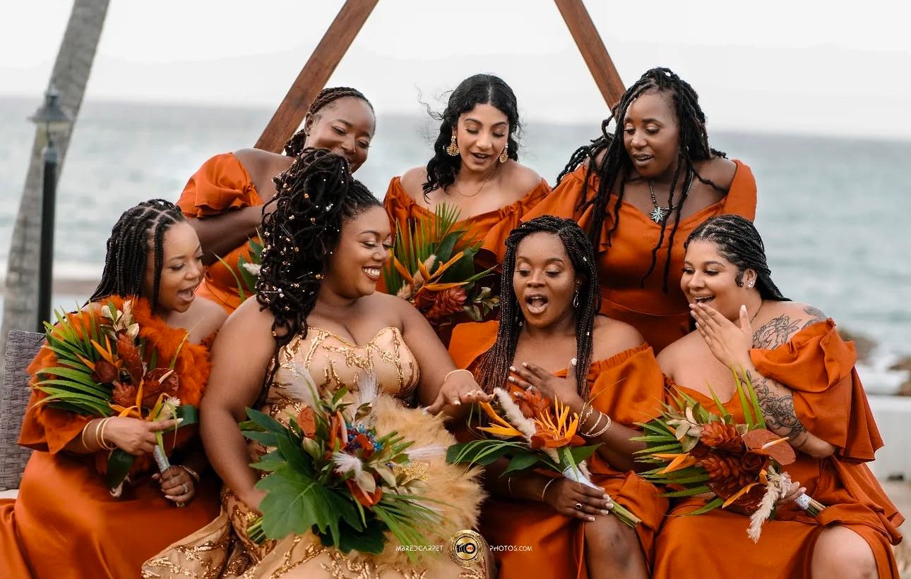 Bridesmaids in vibrant orange dresses celebrate joyfully with tropical flowers.