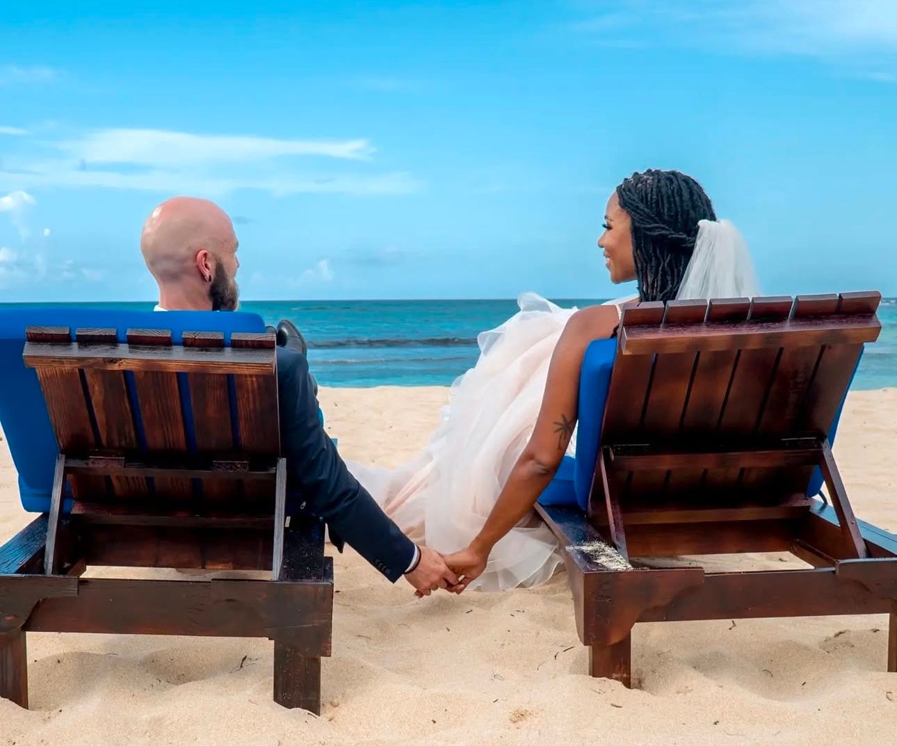 Couple relaxing in beach chairs by the ocean holding hands on a sunny day.