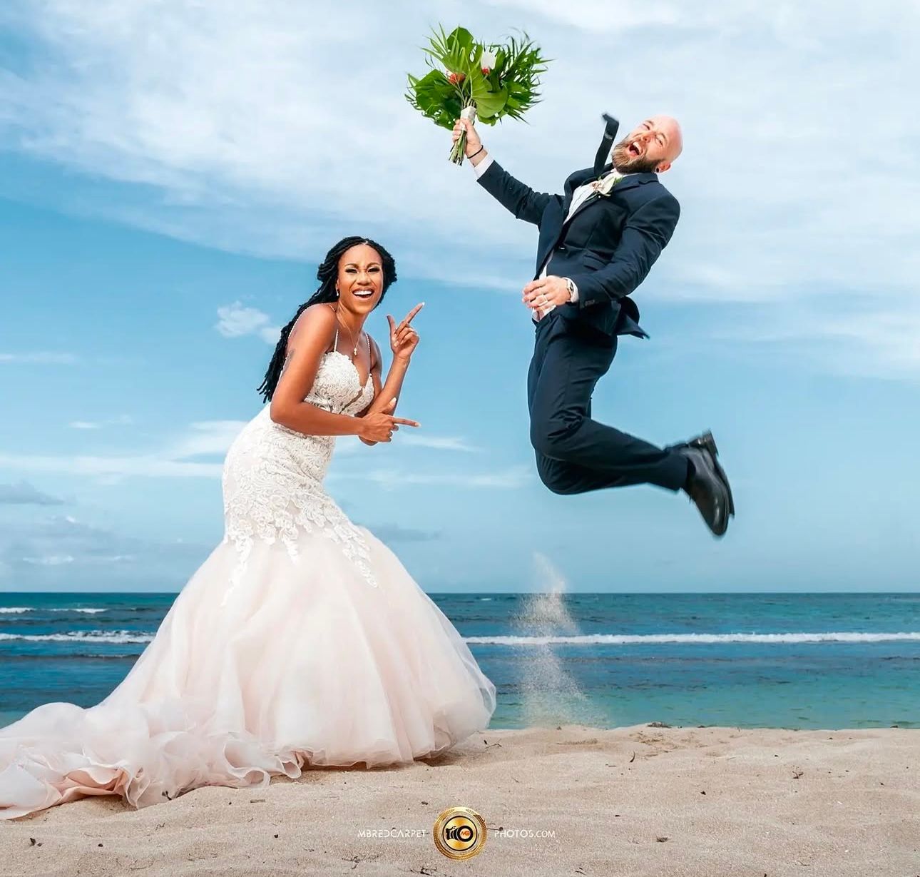 Playful wedding photo with couple jumping on beach, groom holding greenery.