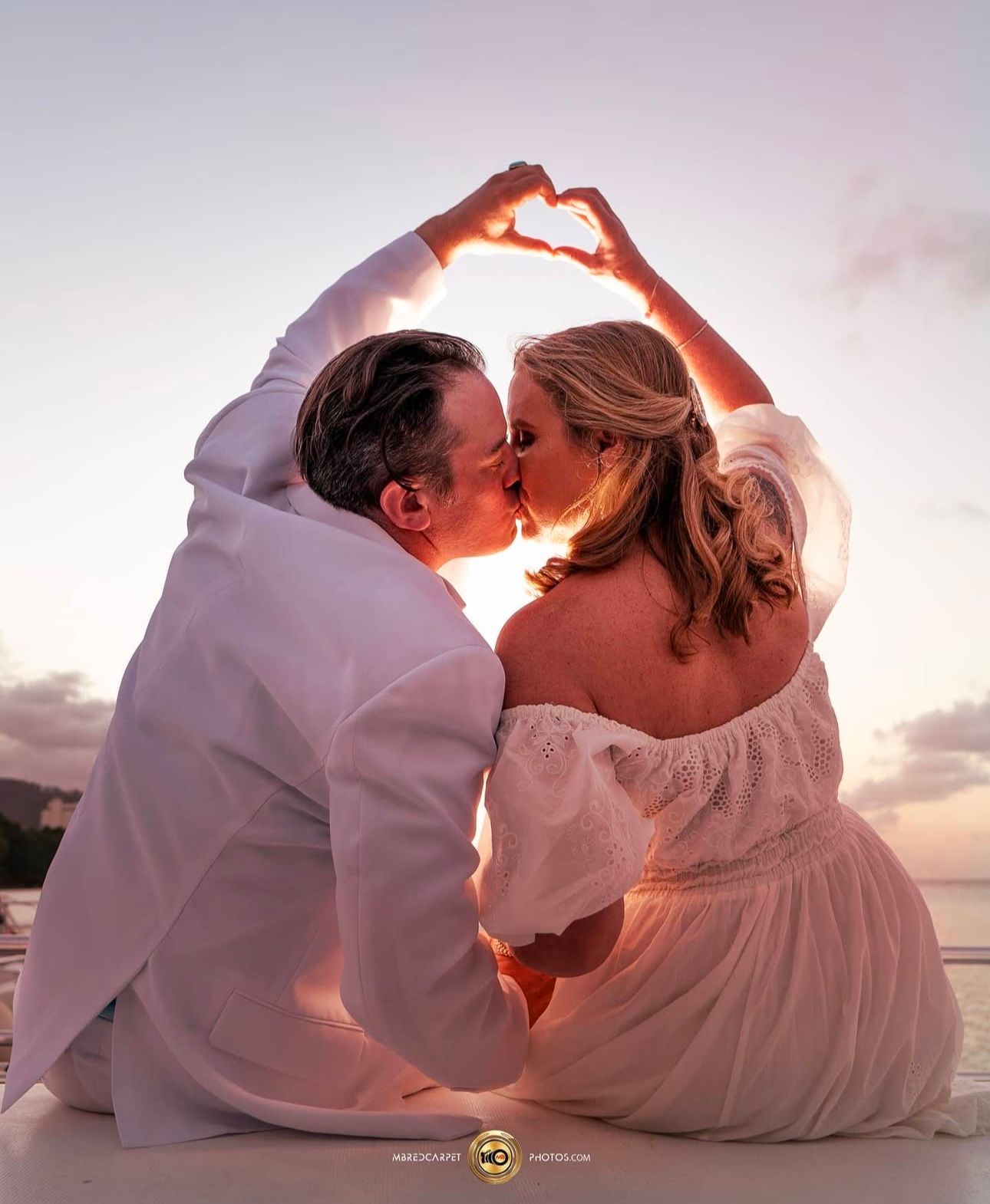 Couple forms heart shape with hands while sharing romantic sunset kiss.