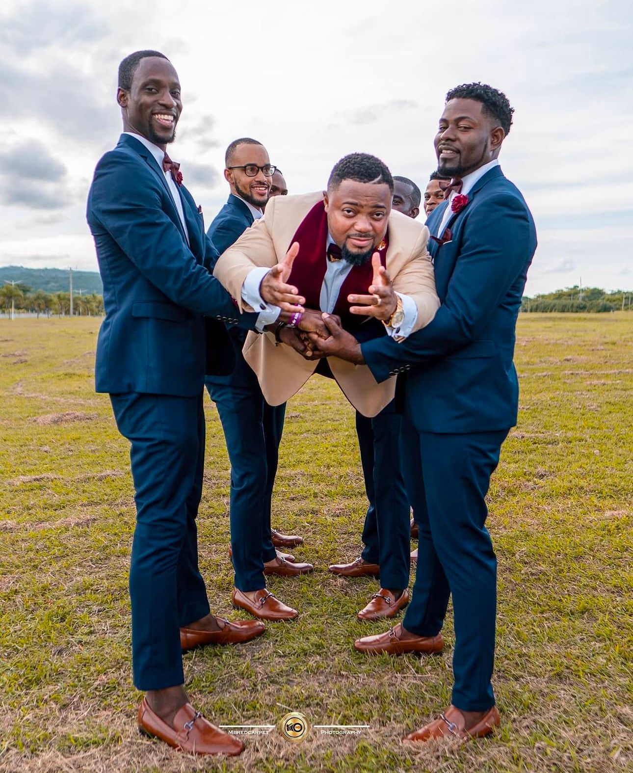 Groomsmen in blue suits share laughs while checking phone in outdoor field setting.