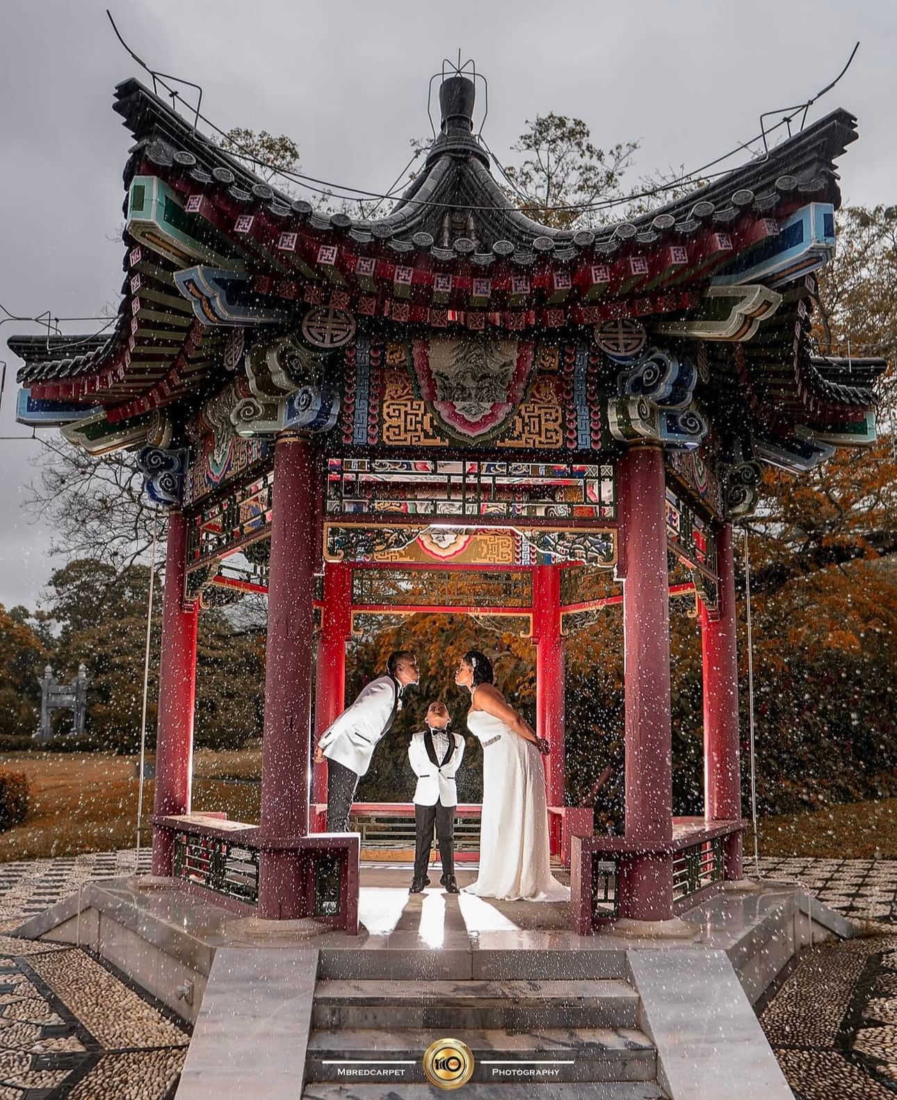 Wedding couple poses under ornate red Chinese pagoda in autumn garden setting.