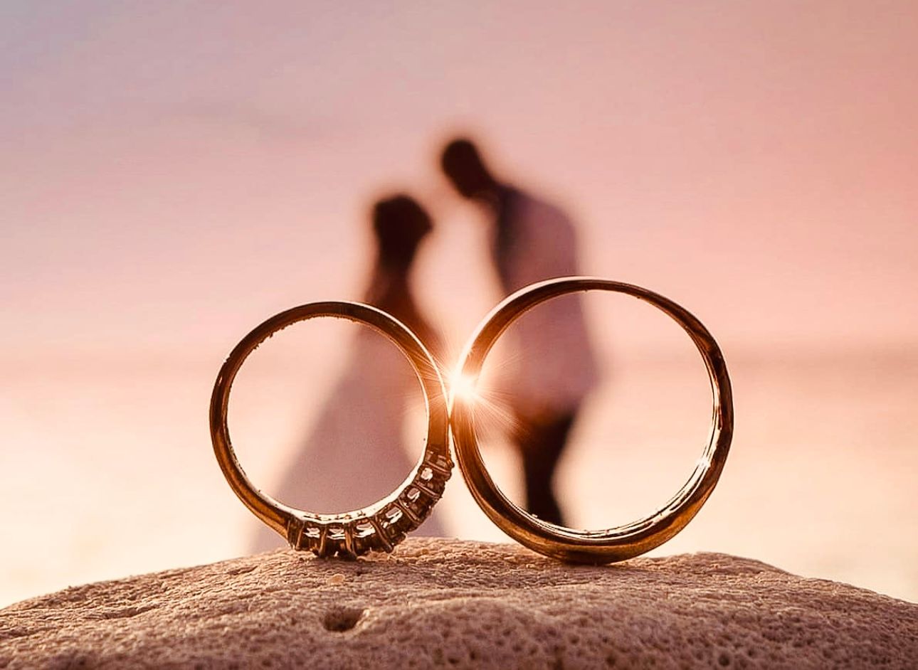 Wedding rings photographed in silhouette against a soft pink sunset background with sand texture.