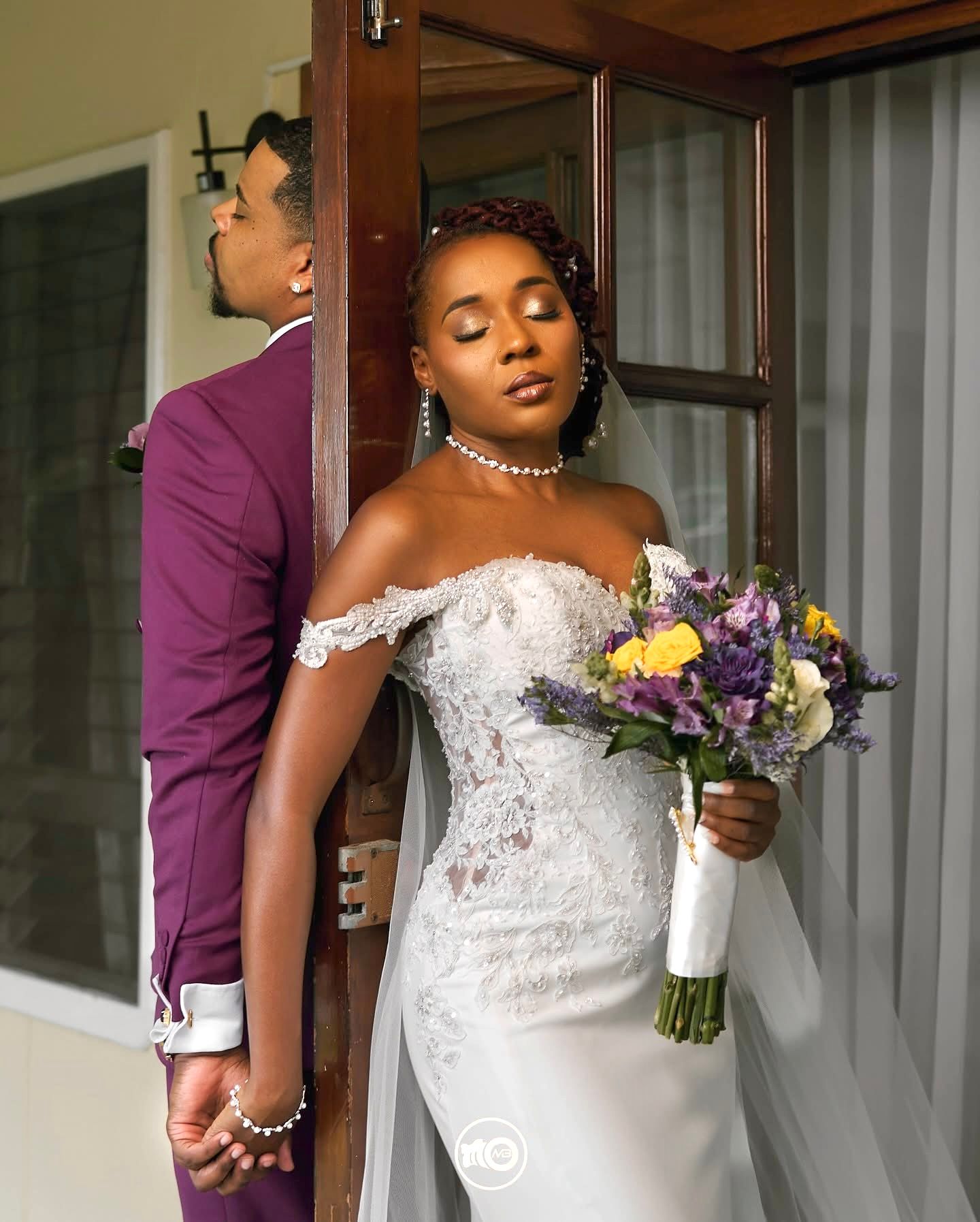 Bride in elegant lace dress holding purple and white floral bouquet.
