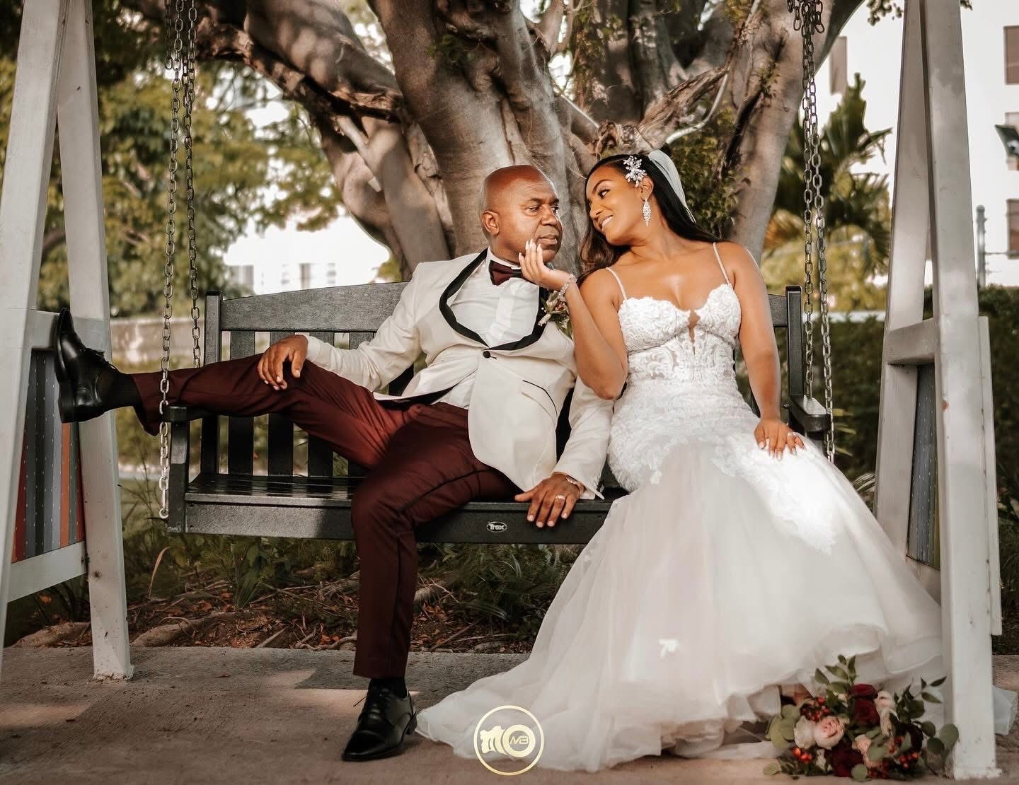 A joyful couple in wedding attire share a playful moment on a swing in an outdoor garden setting.