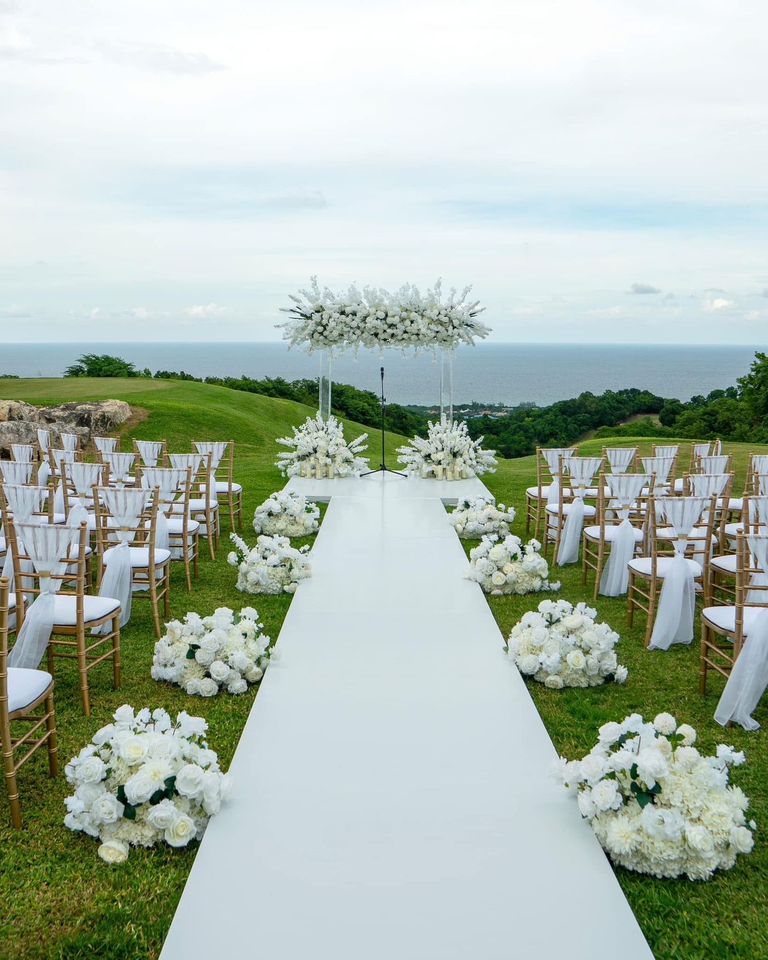 A white wedding aisle runner stretches across a hilltop lawn with chairs and floral arrangements for an outdoor ceremony.
