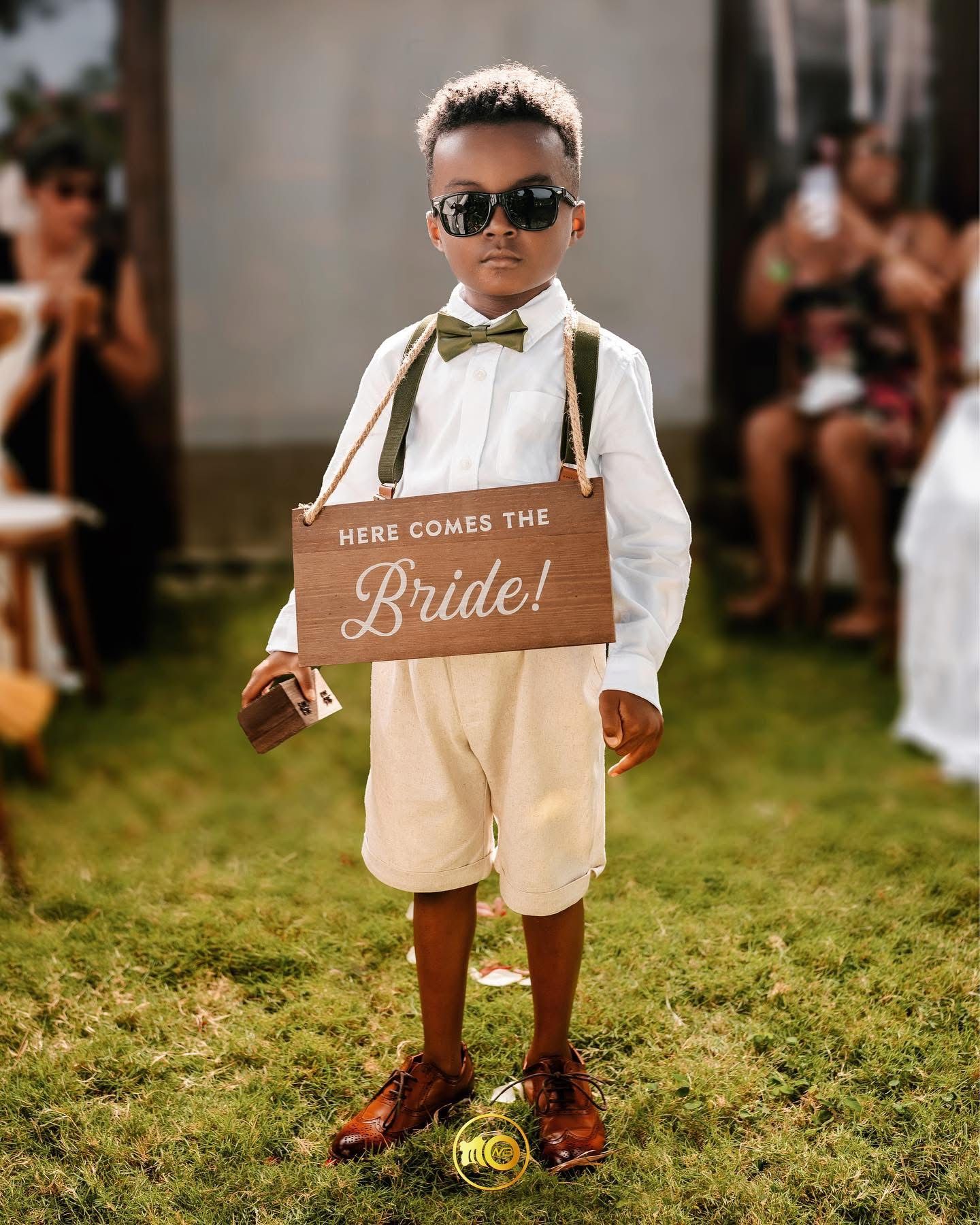 A young ring bearer in suspenders and sunglasses holds a rustic wooden 'Here comes the Bride' sign at an outdoor wedding.