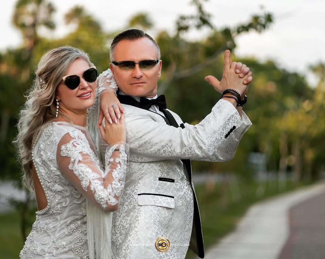 A couple in wedding attire dances together on a tree-lined path during their outdoor celebration.