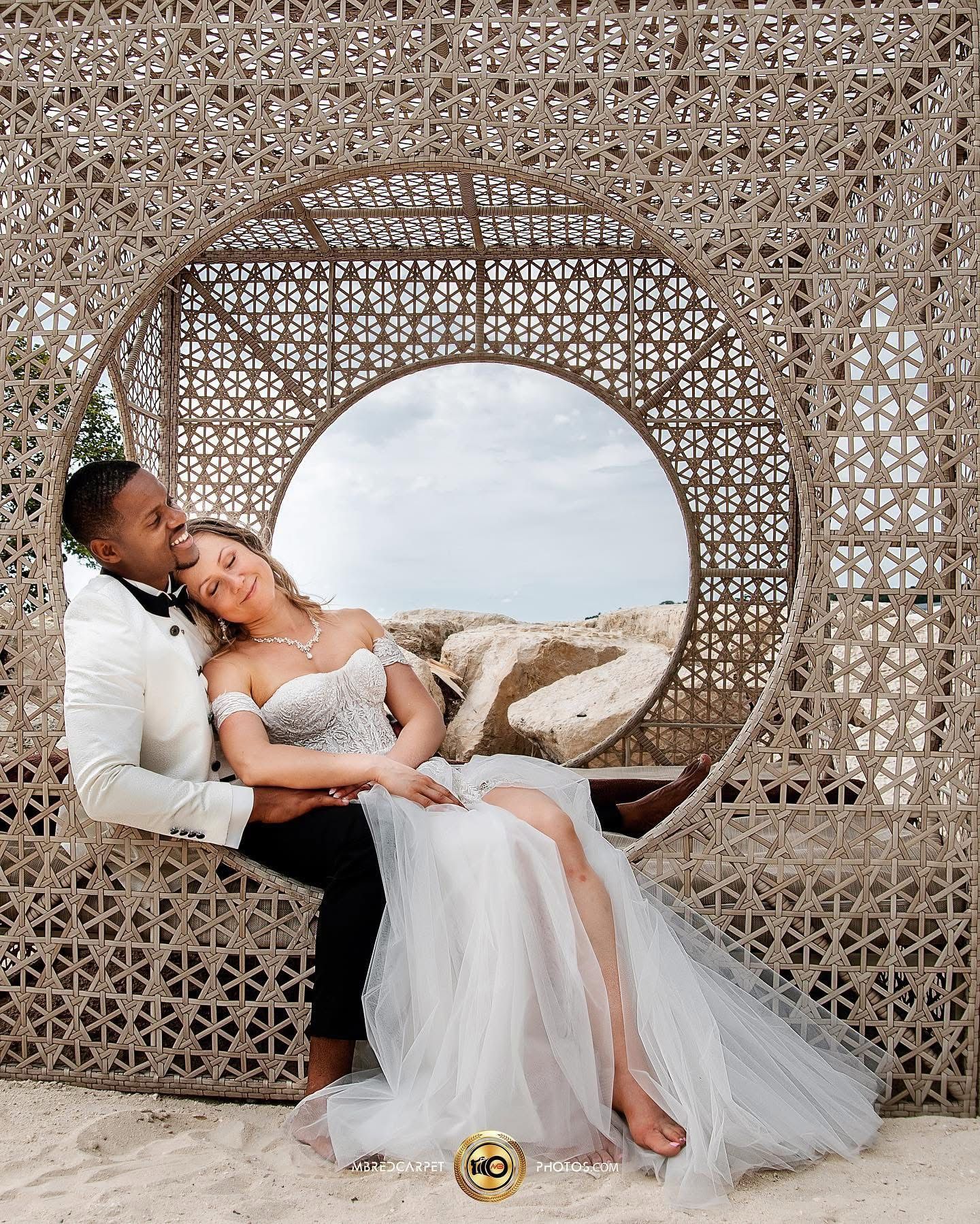 A bride in a white tulle gown reclines on a woven rattan bench against a decorative lattice backdrop.