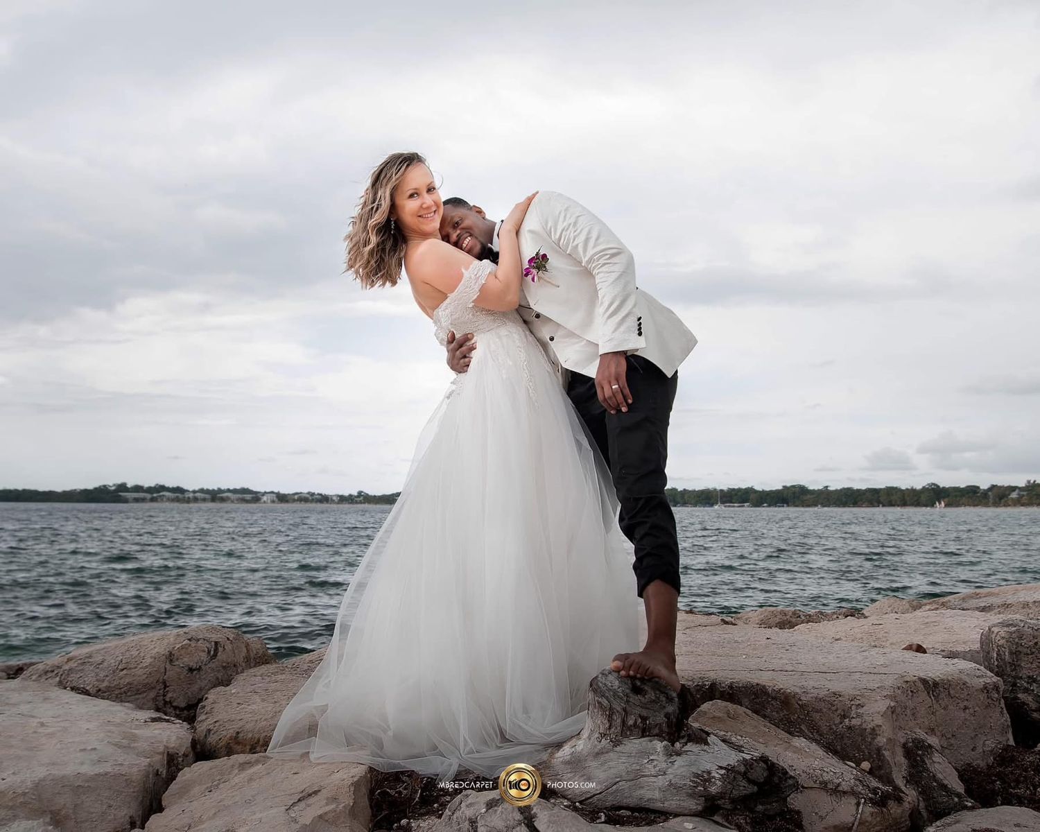 A romantic wedding couple embraces on rocky coastal shores with water and cloudy skies in the background.