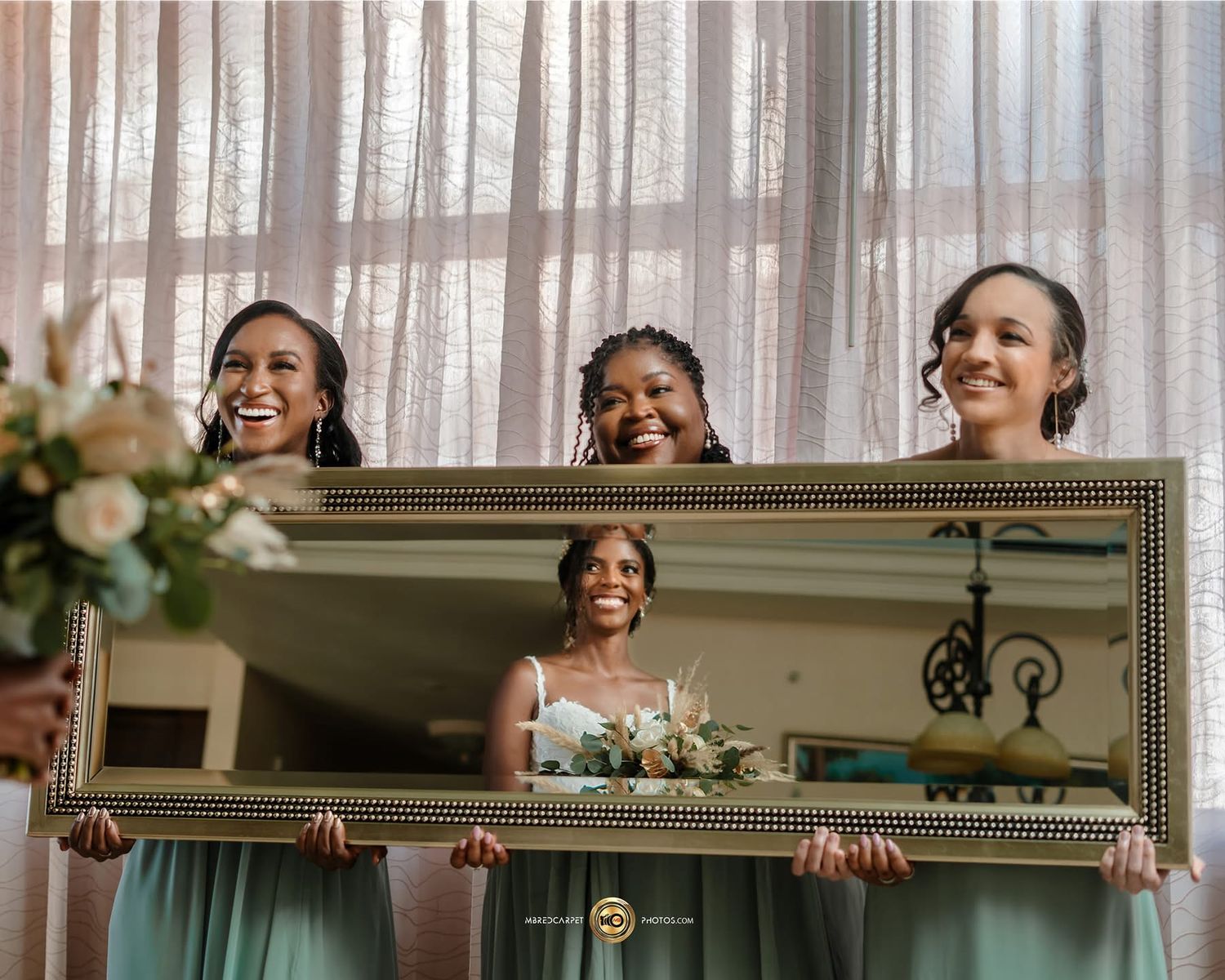 A group of bridesmaids in mint green dresses hold up a large decorative mirror while laughing together.