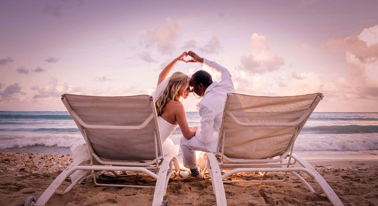 A couple sits on beach chairs making a heart shape with their arms during a romantic sunset by the ocean.