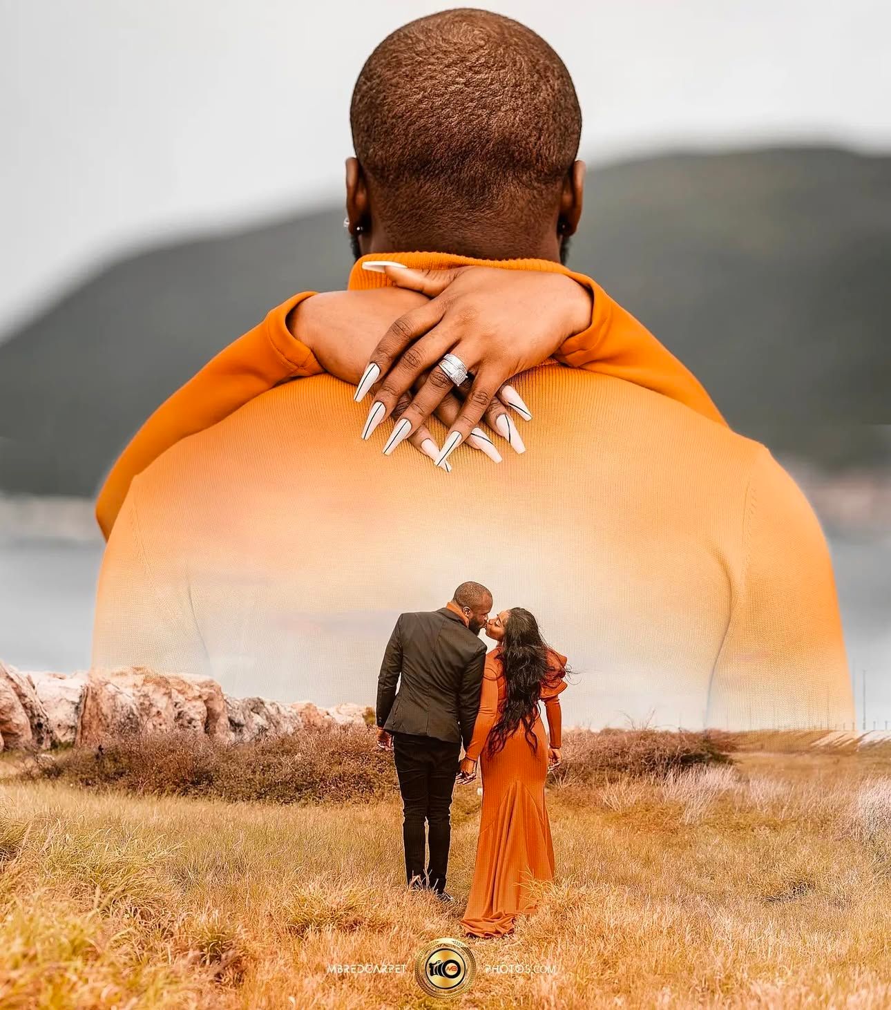 A couple in an orange dress and dark suit stand together in a golden field during sunset with mountains in the background.