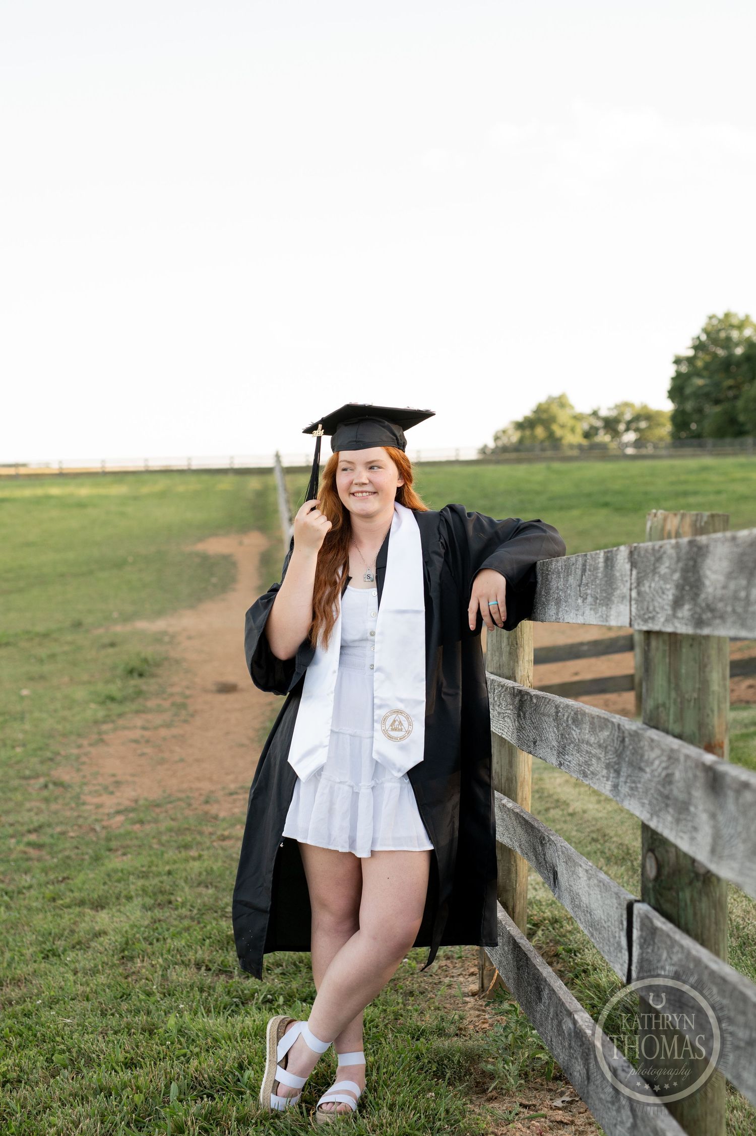 Graduate in white dress and black cap and gown poses by rustic wooden fence in countryside setting.