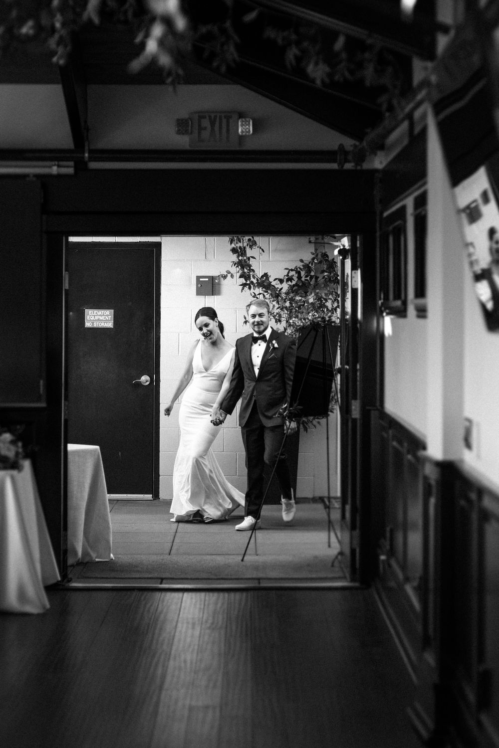 A newlywed couple walks through a dimly lit hallway in formal wedding attire.