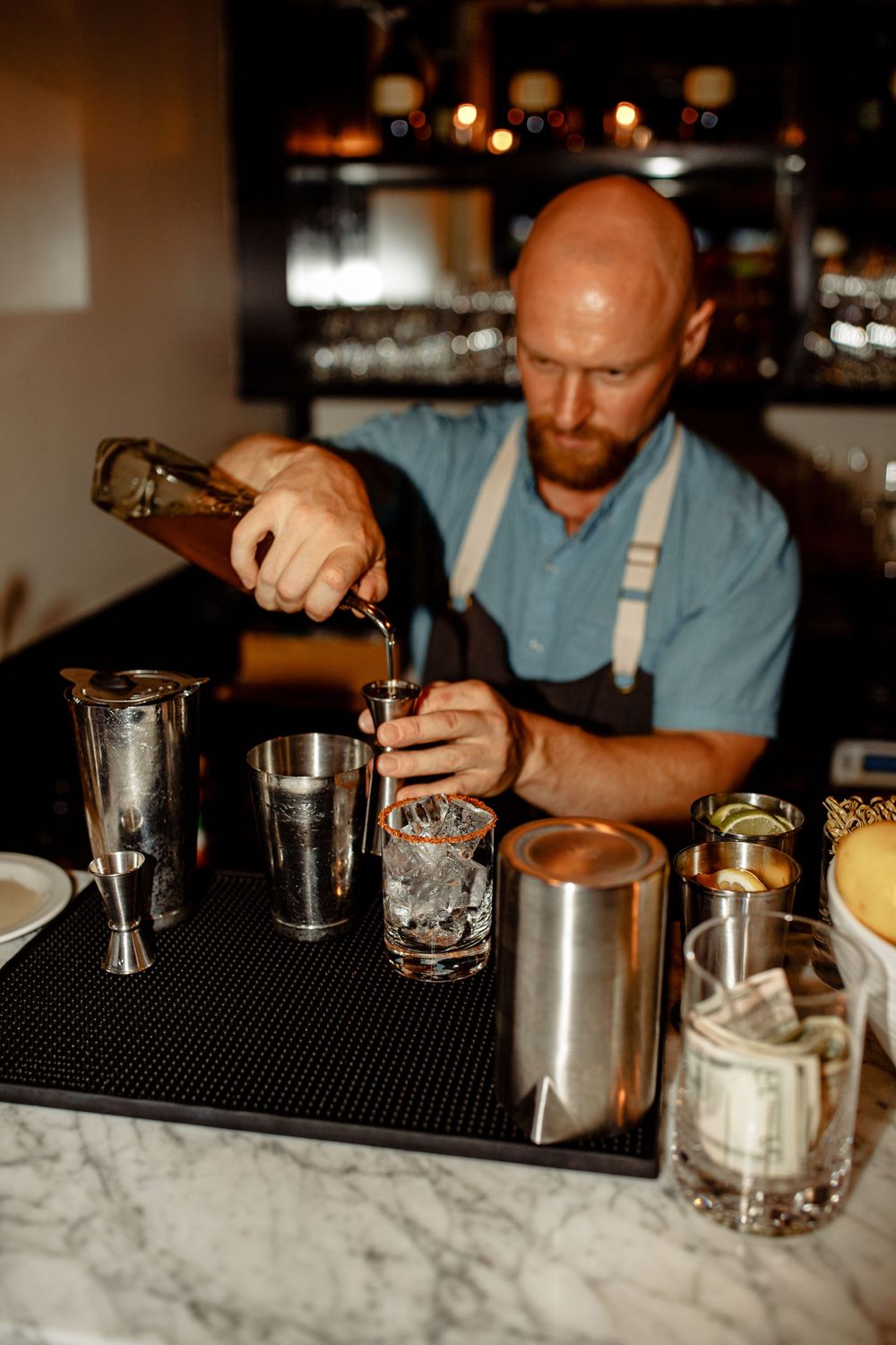 Bartender in suspenders carefully pouring craft cocktail at marble bar counter.