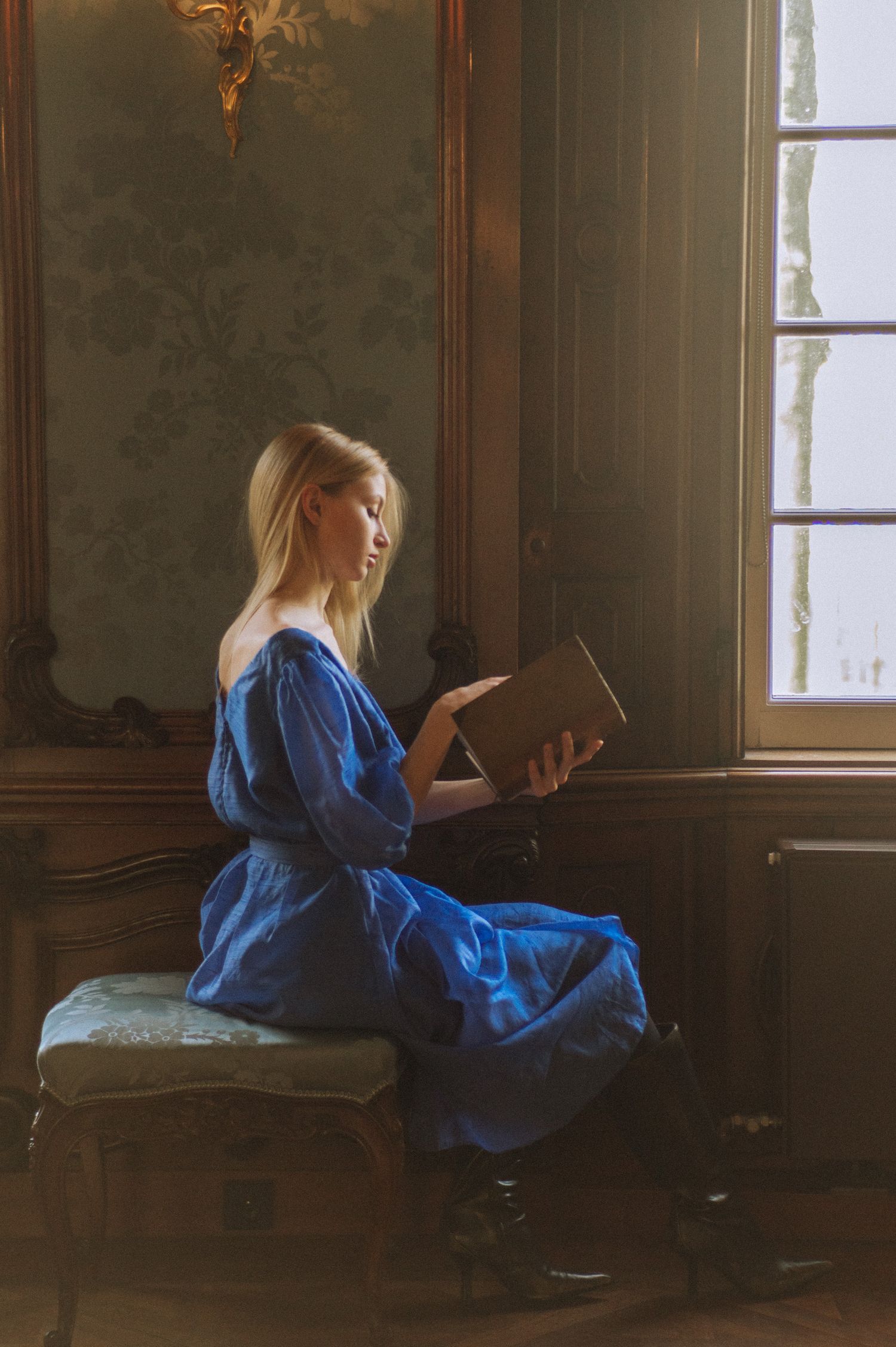 A woman in a blue dress reads by a window in an elegant vintage room.