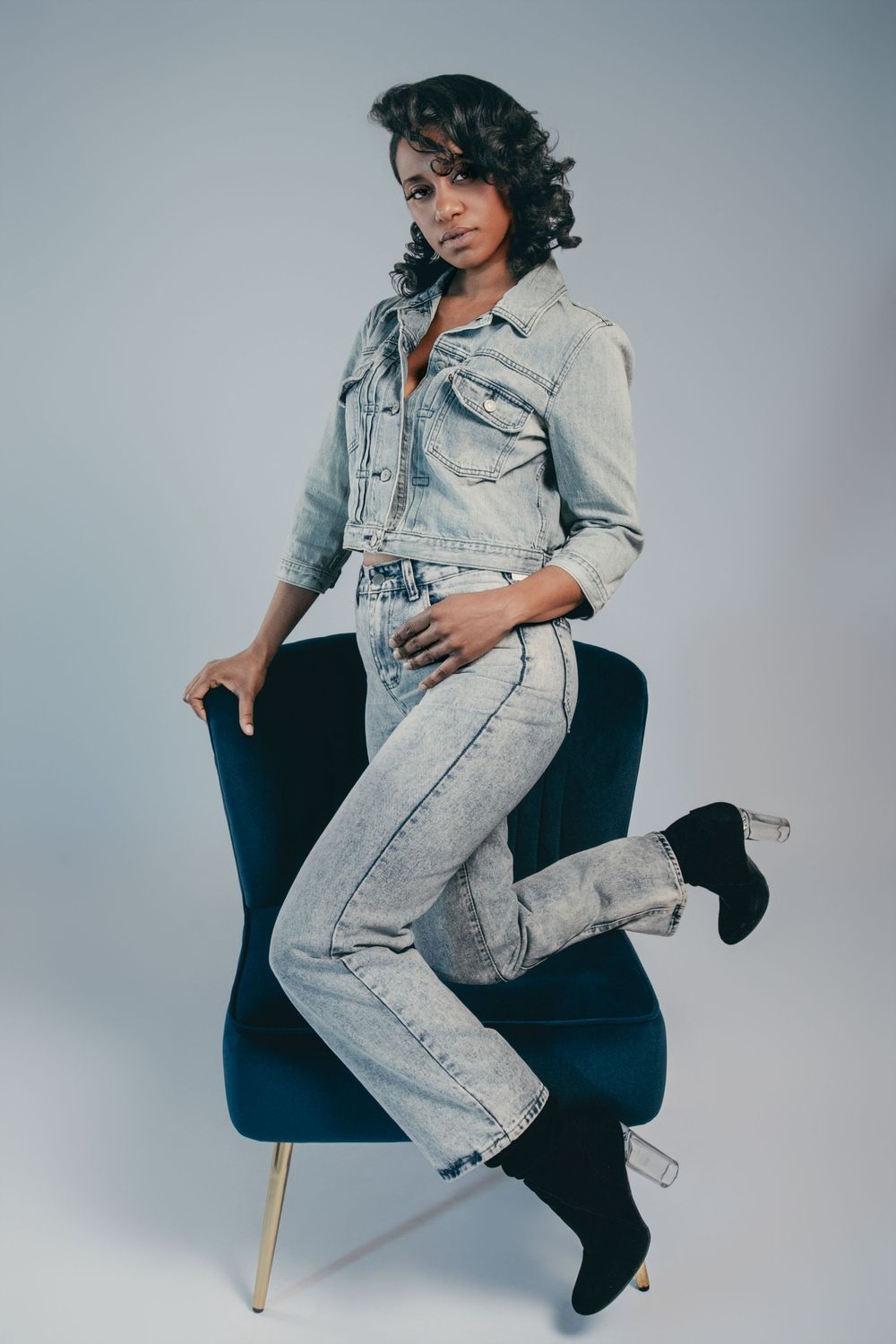 Model in acid wash denim outfit poses artistically on dark blue chair.