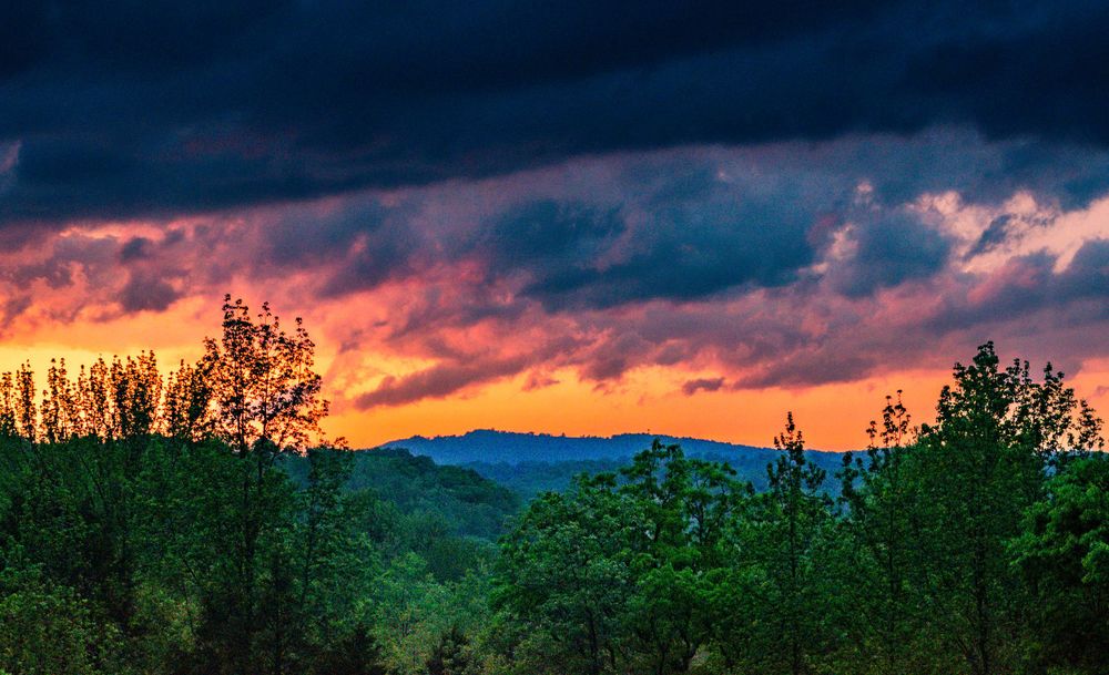Dramatic sunset sky with vibrant orange clouds above silhouetted mountain forest landscape.
