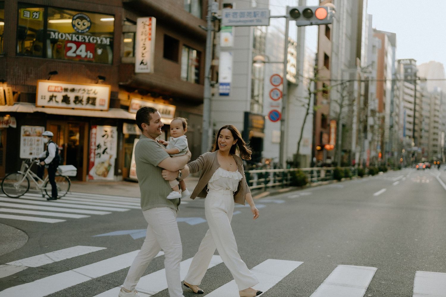 Two people in white outfits cross a city street intersection in Japan with urban buildings and shops visible in background.