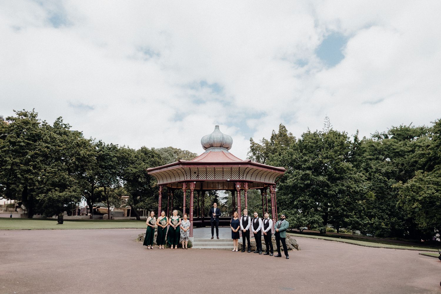 A wedding party gathers in front of an ornate gazebo in a park, captured by Apertured Media Auckland Wedding Photographer.