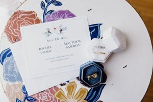 Wedding invitation with floral design and rings in navy velvet box on white table.
