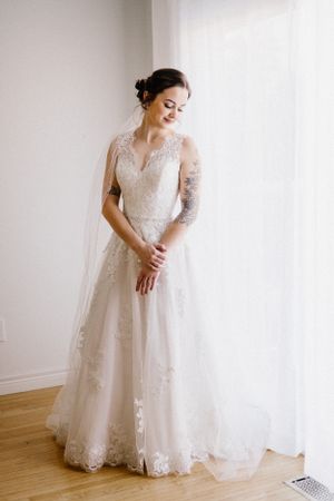 Full length view of bride in delicate lace wedding gown standing by window.