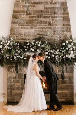 A romantic winter wedding ceremony in front of a stone fireplace adorned with lush white floral arrangements.