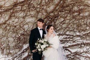 A winter wedding portrait against a rustic wall covered in bare vines.