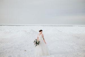 Bride in white gown holding bouquet on snow-covered beach with moody sky.