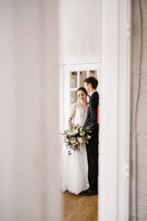 A romantic wedding portrait of a couple in a white dress and black suit embracing by a window.