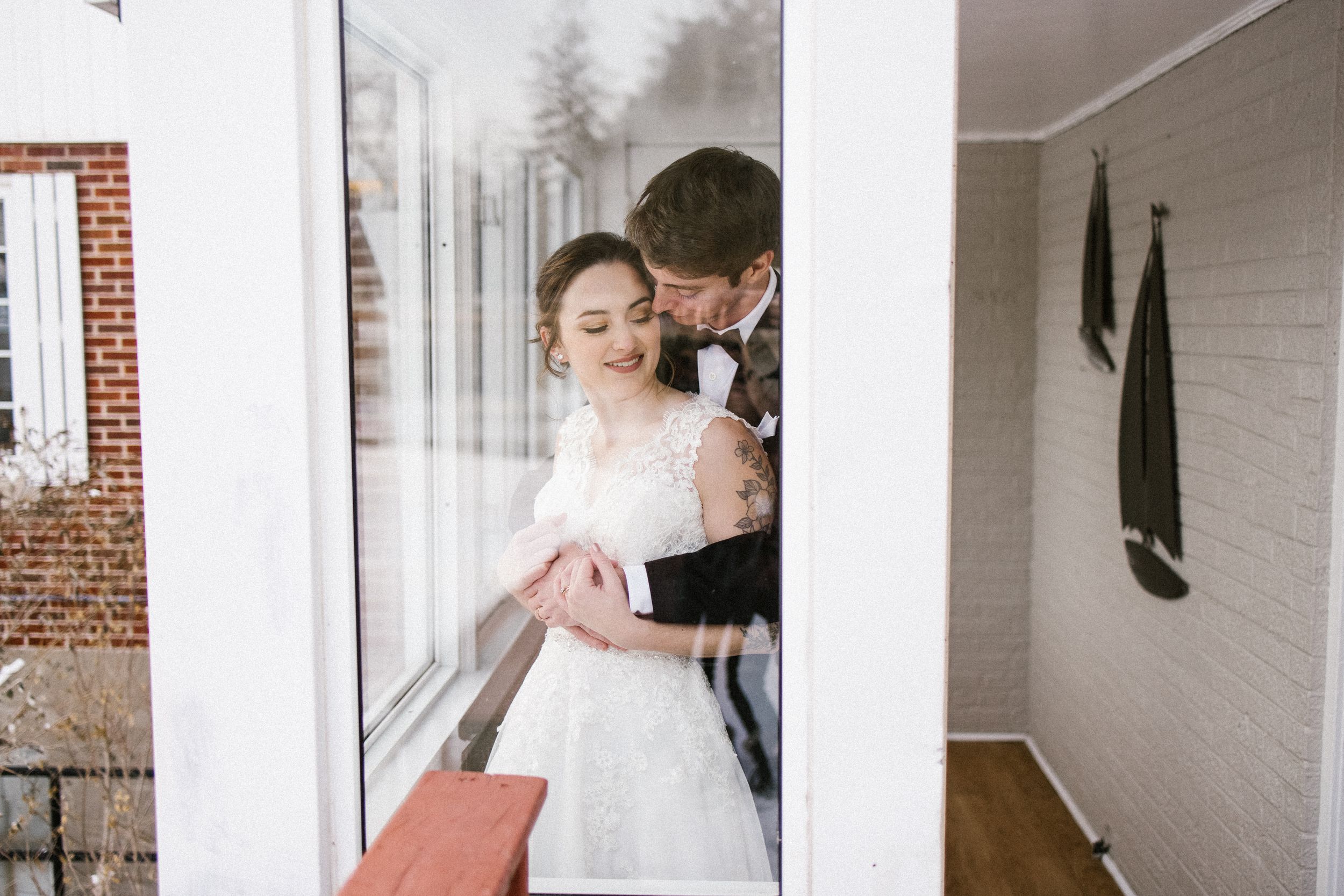A tender moment between newlyweds standing by a window in an intimate indoor setting.