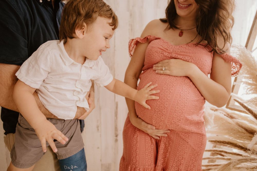 A young child in a white shirt reacts excitedly next to someone wearing a coral-colored dress during a pregnancy photoshoot.