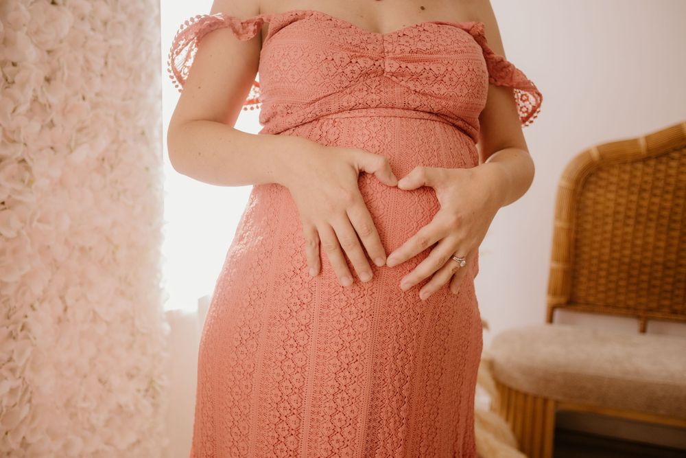 A person wearing a pink knitted dress stands in a room with a wicker chair and white walls.