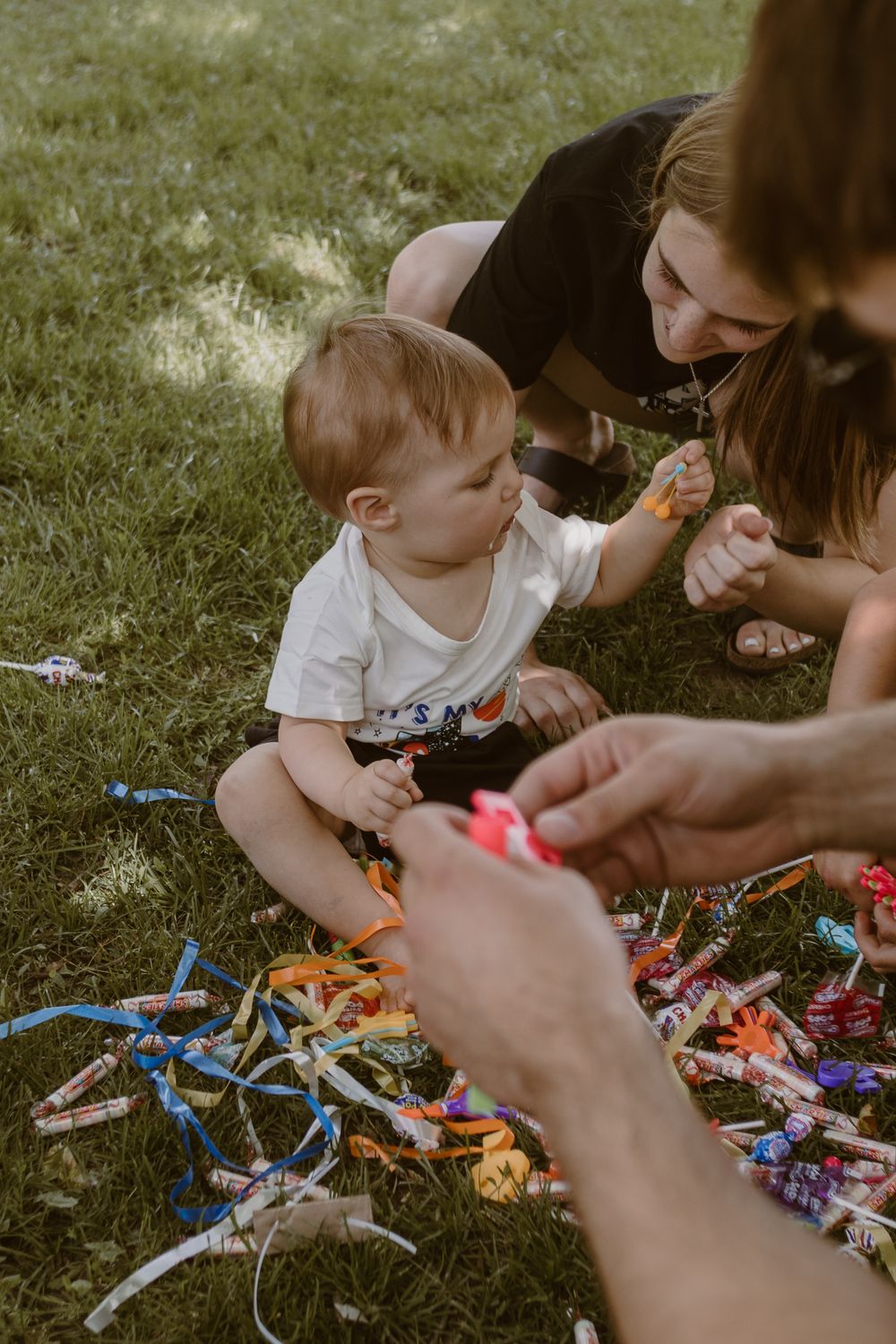 A baby in a white shirt plays with colorful ribbons and party decorations while sitting on grass outdoors.