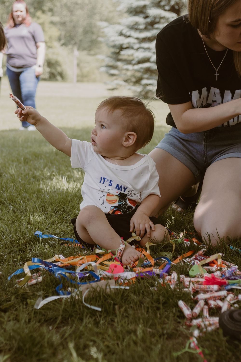 A small child in a white shirt reaches for candy and treats spread on green grass.