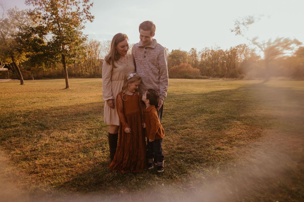 A couple stands together in a park at sunset with golden sunlight filtering through autumn trees in the background.