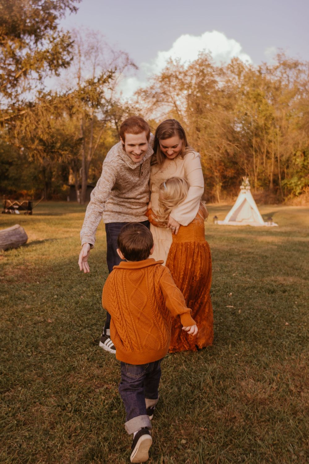 A couple in autumn attire playfully runs through a grassy field with a teepee tent visible in the background.