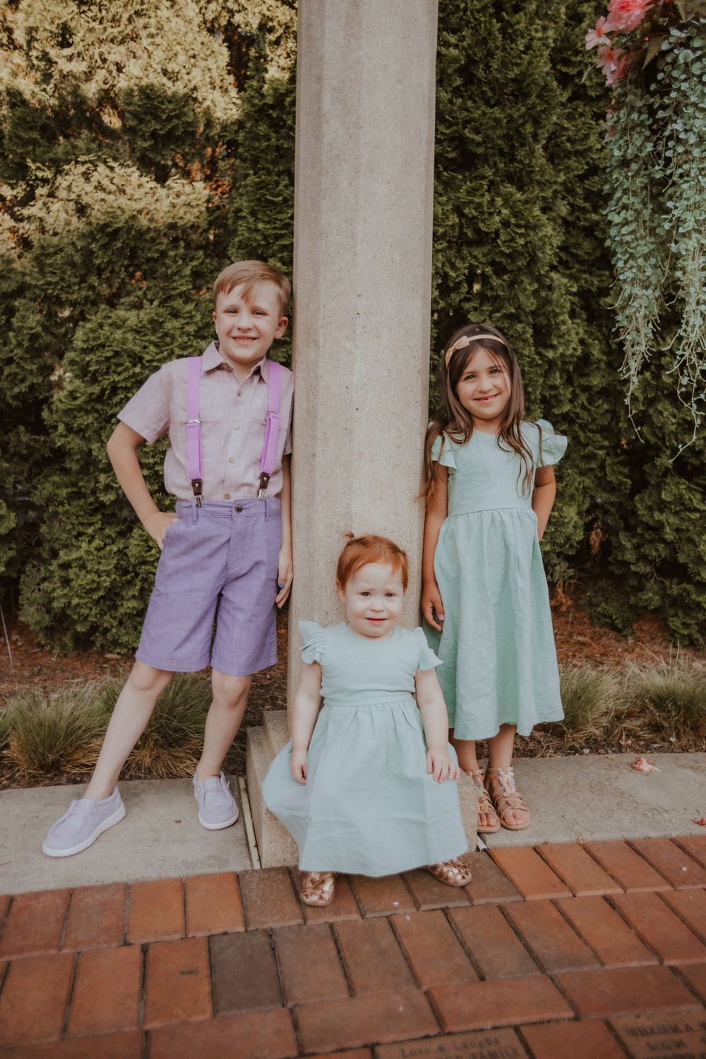 Three children in vintage-inspired outfits pose against greenery with a boy in purple shorts and two girls in mint dresses.