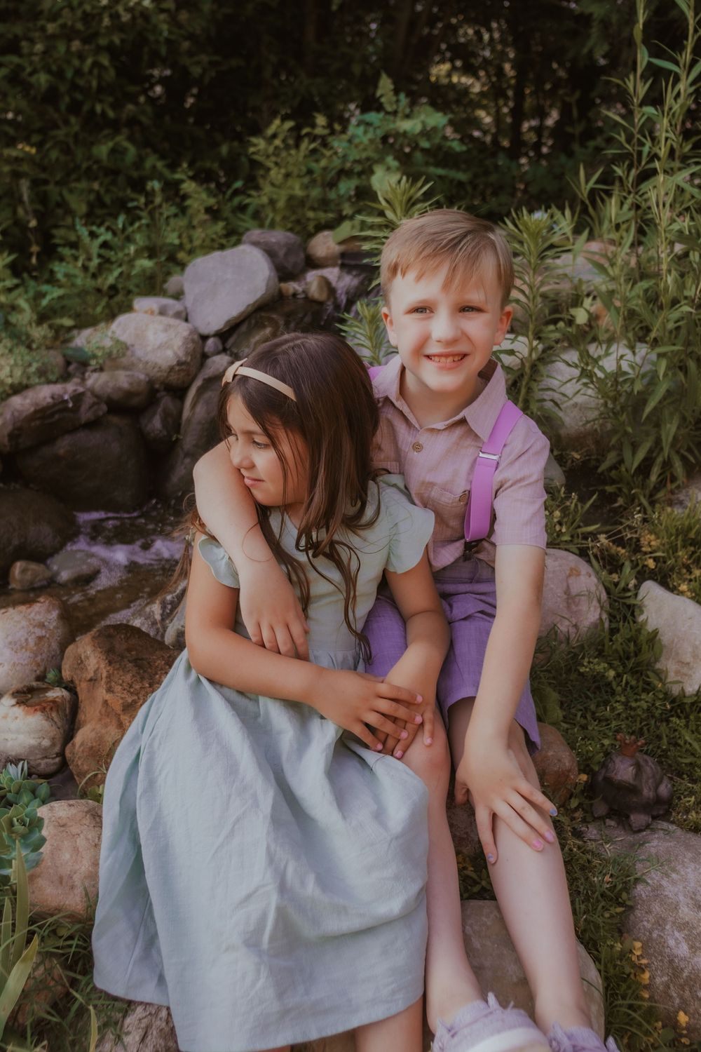 Two children sitting together on rocks surrounded by greenery in a garden setting during summer.