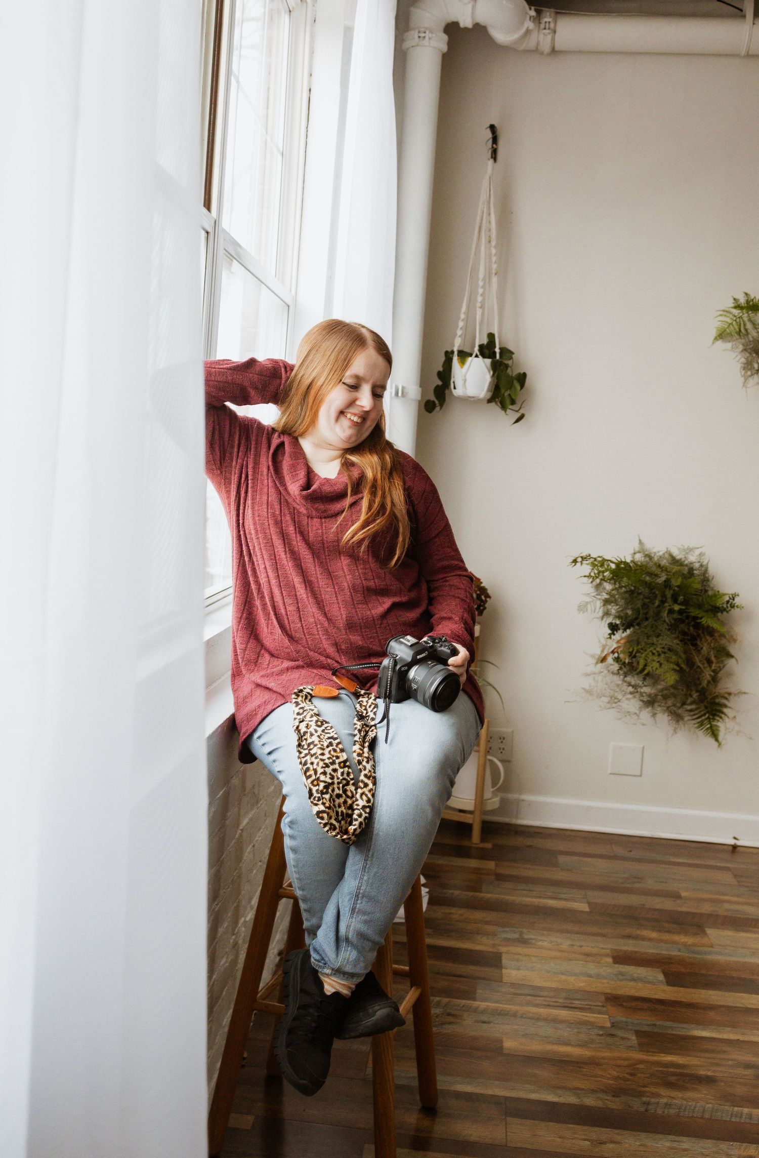 A photographer relaxing by a bright window in a red sweater and jeans while holding a camera.