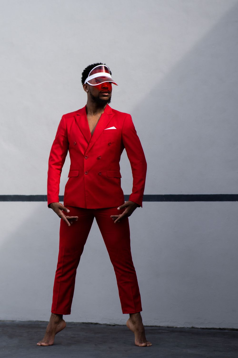 Striking pose in tailored red suit and visor against industrial grey backdrop.