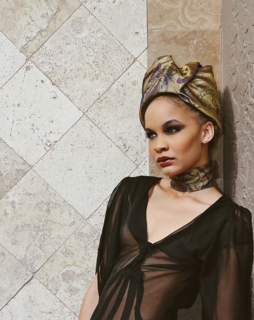 Fashion portrait featuring sheer black top and metallic headpiece against stone wall.