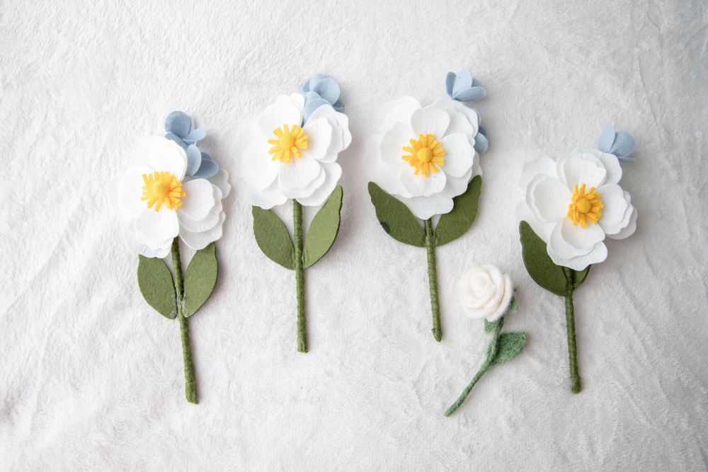 Handmade felt white flower stems with blue and green leaves arranged on white background.